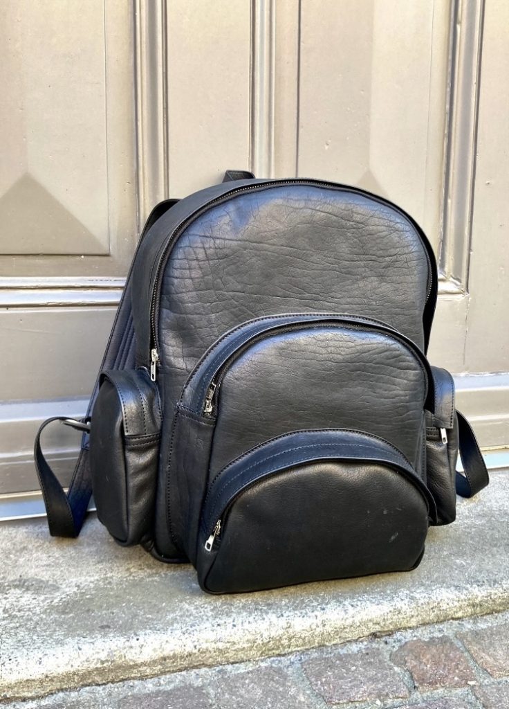 Der große schwarze Lederrucksack mit mehreren Reißverschlussfächern hebt sich deutlich von der hellen Holztüre ab.